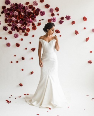 Off the shoulder form-fitting gown in white against a backdrop of flowers.